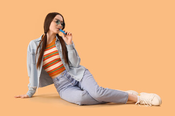 Young woman in sunglasses with disposable electronic cigarette sitting on beige background