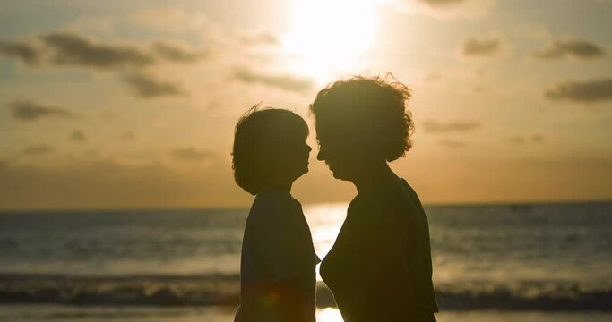 Silhouette of mother and child on orange-yellow sunset, which cutely rub their noses like little fawns, expressing love. Mother and son rub their noses on ocean at sunset. Mother's love for her son.