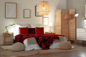 Interior of bedroom with red checkered blankets on bed, blank frames and glowing lamps late in evening