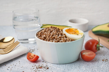 Bowl of tasty buckwheat porridge with soft boiled egg and fresh vegetables on grey table