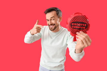Handsome man pointing at whoopee cushion on red background. April Fools' Day celebration
