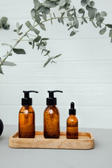 Amber bottles with facial cosmetics on a tray with a branch of eucalyptus on the gray wooden background, facial cleaner, serum. Front view