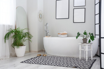 Interior of light bathroom with bathtub, blank frames and houseplants