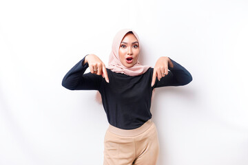 Shocked Asian Muslim woman wearing hijab pointing at the copy space below her, isolated by white...