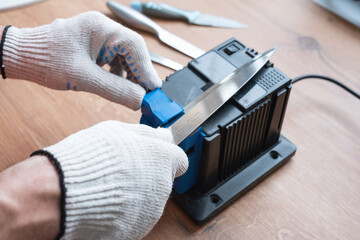 Sharpening a knife on an electric sharpener at home. The man's hand drives the knife blade between the blue sharpeners, dust flies on the machine.
