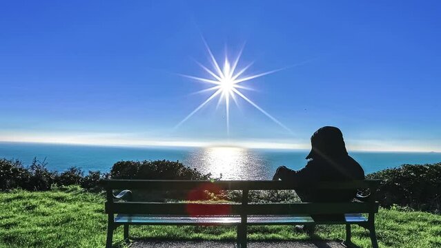 Young man on a park bench watching the ocean with the sun reflecting off the water - siding time lapse