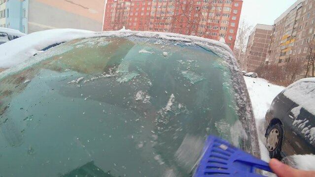 First-person person cleans ice window on car with ice scraper. Focus on ice scraper. Cold snowy and frosty morning. Action Camera Live Camera. Against background of city and dense building houses.