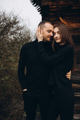 a man and a woman in black clothes near an old wooden house. photo in dark brown tones. old abandoned house and cloudy cold weather. beautiful stylish couple on the background of a brown wooden wall.