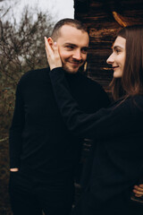 a man and a woman in black clothes near an old wooden house. photo in dark brown tones. old abandoned house and cloudy cold weather. beautiful stylish couple on the background of a brown wooden wall.
