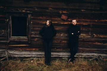 a man and a woman in black clothes near an old wooden house. photo in dark brown tones. old abandoned house and cloudy cold weather. beautiful stylish couple on the background of a brown wooden wall.