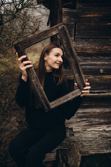 beautiful girl in black clothes near an old wooden house, holding a wooden window frame in her hands. war in Ukraine
