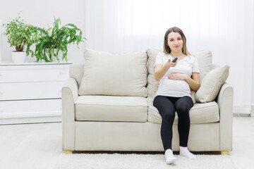 Obraz premium Pregnant woman sitting on sofa with remote control.