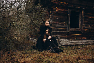 a man and a woman in black clothes sit near an old wooden house. photo in dark brown tones. old abandoned house and cloudy cold weather. hug a loved one