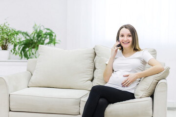 Fototapeta premium Smiling pregnant woman talking over the phone on white sofa.