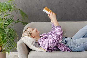 Side view young fun smart woman wearing casual clothes reading book novel lying on grey sofa couch...