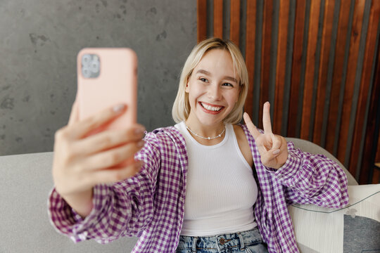 Young woman wear casual clothes do selfie shot on mobile cell phone show v-sign sits on grey sofa couch stay at home hotel flat rest relax spend free time in living room indoor. People lounge concept.