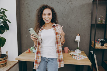 Young fun smart satisfied student successful employee business woman of African American ethnicity wearing casual shirt sit on table use mobile cell phone showing thumb up work at office desk indoors.