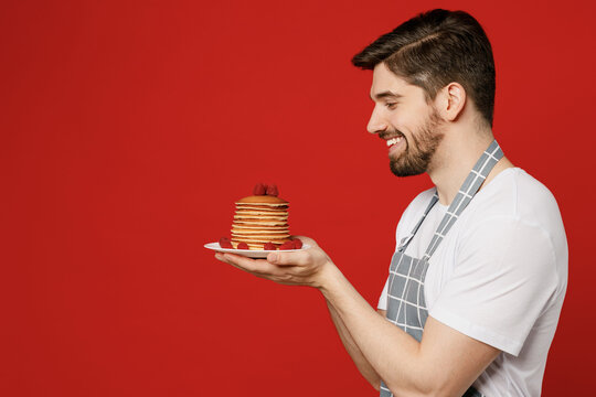 Side View Young Cheerful Fun Male Housewife Housekeeper Chef Cook Baker Man Wears Grey Apron Hold In Hand Plate With Pancakes Look Camera Isolated On Plain Red Background Studio. Cooking Food Concept.