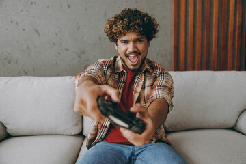 Young Indian man wears casual clothes hold in hand play pc game with joystick console sits on grey...