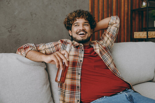 Young smiling fun Indian man wears casual clothes hold bottle of beer look aside sits on grey sofa couch stay at home hotel flat rest relax spend free spare time in living room indoor. Lounge concept. - Powered by Adobe