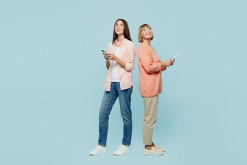 Full body fun happy elder parent mom with young adult daughter two women together wear casual clothes hold in hand mobile cell phone look overhead isolated on plain blue background Family day concept