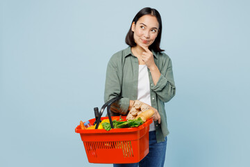 Plakat Young minded woman wears casual clothes hold red basket with food products for preparing dinner look aside isolated on plain blue background studio portrait. Delivery service from shop or restaurant.