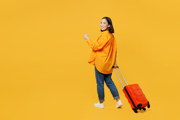 Back view young woman wear summer clothes walk go with suitcase bag show thumb up isolated on plain yellow background. Tourist travel abroad in free time rest getaway. Air flight trip journey concept.