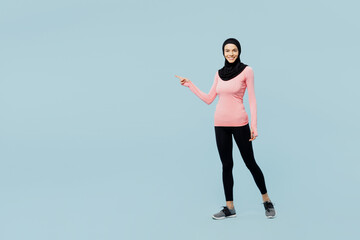 Full body fun young asian muslim fitness trainer sporty woman wear pink abaya hijab spend time in home gym point finger aside isolated on plain blue background studio. Workout sport fit abs concept.