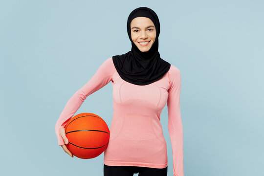 Young happy arabian asian muslim fitness trainer sporty woman wear pink abaya hijab spend time in home gym hold basketball ball isolated on plain blue background studio Workout sport fit abs concept. - Powered by Adobe