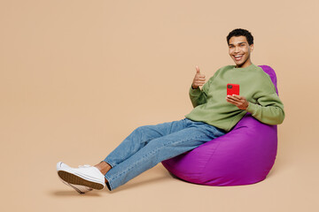 Full body satisfied fun young man of African American ethnicity wear green sweatshirt sit in bag chair hold in hand use mobile cell phone show thumb up isolated on plain pastel light beige background.