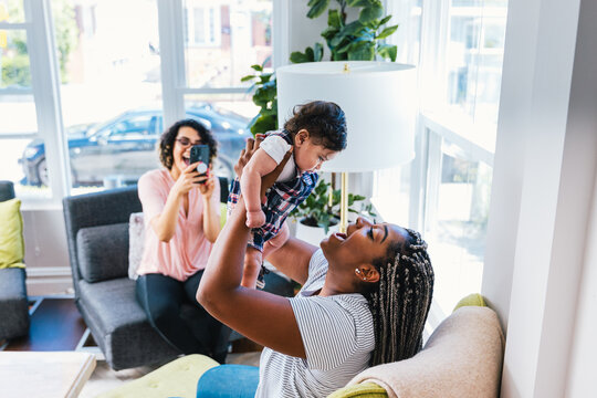 Happy mother playing with cute son while girlfriend photographing them in living room