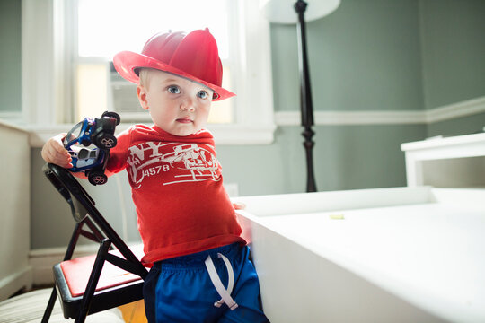 Toddler boy dressed up as fireman stands from chair holding toy car