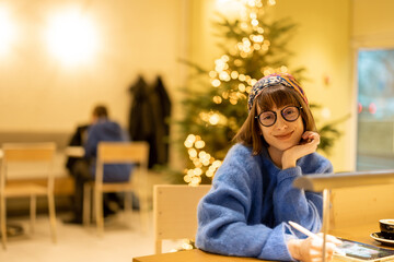 Portrait of young adult woman sitting at modern coffee shop in yellow tones and Cristmas tree on background. Concept of remote creative work online