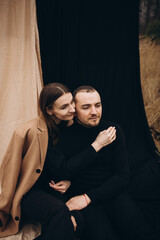 Fototapeta na wymiar a man and a girl in black clothes and a beige coat on a black and beige fabric background. man and woman hugging, cold cloudy weather and dry yellow tall grass . stylish fashion portrait.