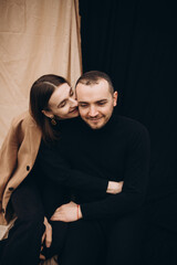 a man and a girl in black clothes and a beige coat on a black and beige fabric background. man and woman hugging, cold cloudy weather and dry yellow tall grass . stylish fashion portrait.