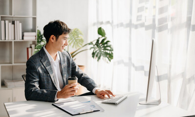 Confident Asian businessman typing laptop computer and digital tablet while holding coffee at modern office