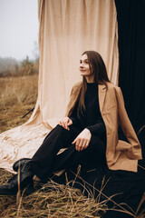 beautiful girl in a black sweater and black pants and a beige coat on a black and beige fabric background. cold cloudy weather and dry yellow tall grass and a girl. stylish fashion portrait. 