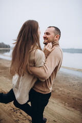 to carry his beloved girl in his arms. a love story of a man and a woman on the shore of a foggy lake. stylish couple in beige sweaters in cold weather on the lake