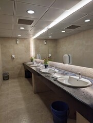 A large washroom with a lot of basins on a row with a big mirror hanging on the wall, a blue...