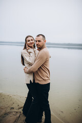 man and woman hugging on the lake shore, cold foggy weather. romantic photo in beige clothes. to...