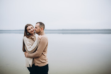 man and woman hugging on the lake shore, cold foggy weather. romantic photo in beige clothes. to...