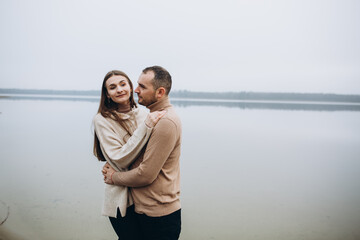 man and woman hugging on the lake shore, cold foggy weather. romantic photo in beige clothes. to hug a woman, a love story. beige sweaters. romantic photos against the background of a foggy lake