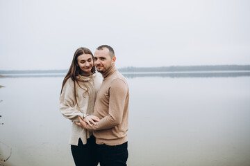 man and woman hugging on the lake shore, cold foggy weather. romantic photo in beige clothes. to...