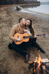 Fototapeta premium a man on the shore of a lake near a fire plays the guitar for a girl. foggy cold weather. beige men's coat. bonfire on the shore of the reservoir. photo in beige tones. romantic photo with a guitar 