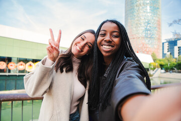 A couple of young multiracial women having fun taking a selfie photo portrait. Front view of one happy african american girl and her friend, a pretty caucasian brunette smiling doing the peace sign