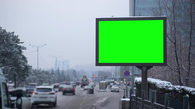 4k Mockup poster stands on street with passing cars in city spbas. Lot of transport raffic is driving on road and green screen lightbox standing outdoors in snowy winter weather. Vertical billboard is