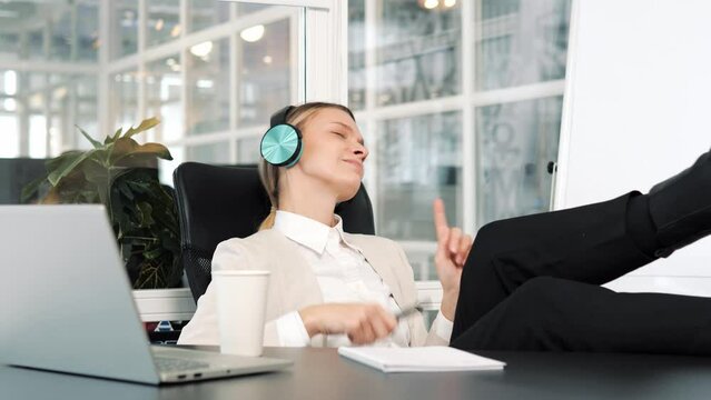 Happy funky businesswoman relaxing wearing wireless green headphones listening to music pretending playing drums at workplace, funny female worker enjoy favorite track having fun during work break.