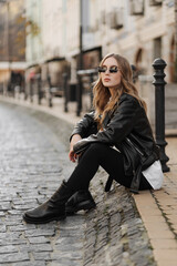 Fashionable  blonde woman model with  black leather jacket and style sunglasses sitting on a floor at the city 