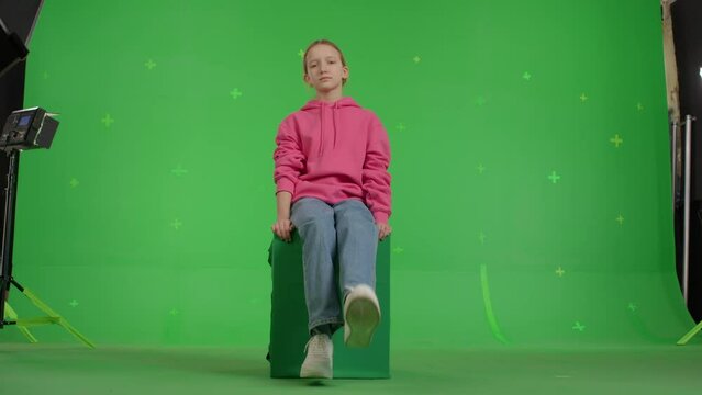 The girl sits on an imaginary parapet or roof. Teenager waving her legs while sitting in a virtual window on green screen background.