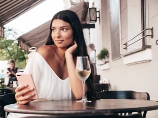 Young beautiful smiling hipster female in white dress. Sexy carefree woman sitting at cafe in the street. Positive model drinking white wine. Cheerful and happy. Holding smartphone